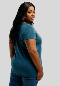 Donna con lunghi capelli neri si gira leggermente, indossando una maglietta turchese e jeans, mostrando un dettagliato tatuaggio di leone sul braccio sinistro superiore, su uno sfondo grigio chiaro.