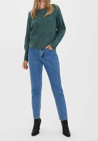 Pull en tricot vert à manches longues, associé à un jean taille haute bleu clair. Le modèle porte des bottines noires. Design simple, tissu texturé.