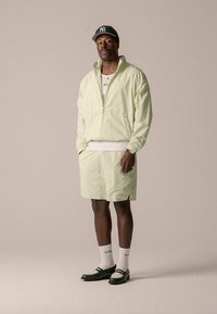 Lichtgroen jack met ritssluiting en een gestructureerd patroon, gecombineerd met bijpassende korte broek. Afgewerkt met zwarte schoenen, witte sokken en een groene pet.