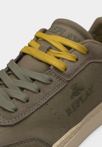Baskets taupe avec des lacets bicolores jaune et vert, textures en daim et tissu, et logo "Replay" en relief sur le côté.