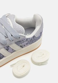 Gray suede sneaker with lavender snake print accents, three white stripes, and white laces. Includes two rolled white shoelaces.