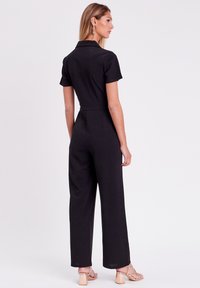 Combinaison noire à manches courtes, avec col, taille cintrée et silhouette évasée. Tissu lisse avec une finition nette et sans motifs visibles.