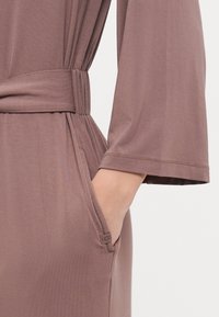 Braunes Stoffkleid mit tailliertem Gürtel, langen Ärmeln und seitlichen Taschen. Glatte Textur mit subtiler Dehnbarkeit für zusätzlichen Komfort.