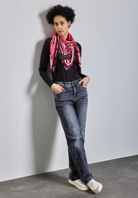 Svart långärmad topp, ljusrosa, svart och vit mönstrad scarf, lätt blekta blå jeans och ljusa sneakers, poserande mot en grå vägg.