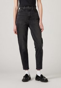 Personne portant un jean noir taille haute, des chaussettes blanches et des mocassins noirs, debout devant un fond clair uni.