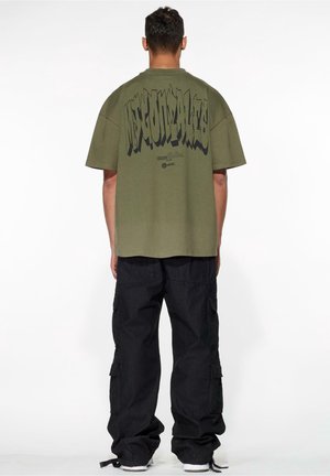 Homme portant un t-shirt oversize vert olive avec un texte noir en gras et un pantalon cargo noir, debout de dos sur fond blanc.