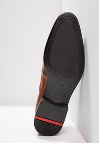 Braune Lederschuhe mit einer soliden schwarzen Gummisohle und einem roten Akzentstreifen. Verfügt über eine strukturierte Verzierung entlang der Sohlenkante.