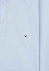 Camisa polo azul claro con cuello, con tres botones y un pequeño logo de tijeras negras en la parte izquierda del pecho. Tejido de algodón suave.