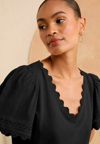 Melna struktūrēta augšdaļa ar īsām pūkām, viļņotu kakla izgriezumu un acu caurumu detaļām pie piedurkņu maliņām.