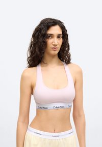 Νεαρή γυναίκα με μακριά σγουρά μαλλιά, φορούσα ανοιχτό ροζ αθλητικό σουτιέν Calvin Klein και ανοιχτόχρωμο κάτω μέρος, μπροστά σε ένα απλό φόντο.