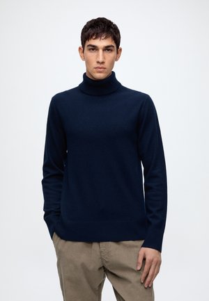 Jeune homme portant un pull à col roulé bleu marine et un pantalon en velours côtelé beige, debout devant un fond clair uni.