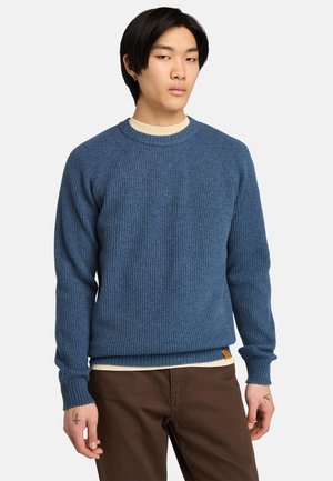 Blauwe geribde trui met een ronde halslijn en lange raglanmouwen, gecombineerd met een crèmekleurig hemd en bruine broek.