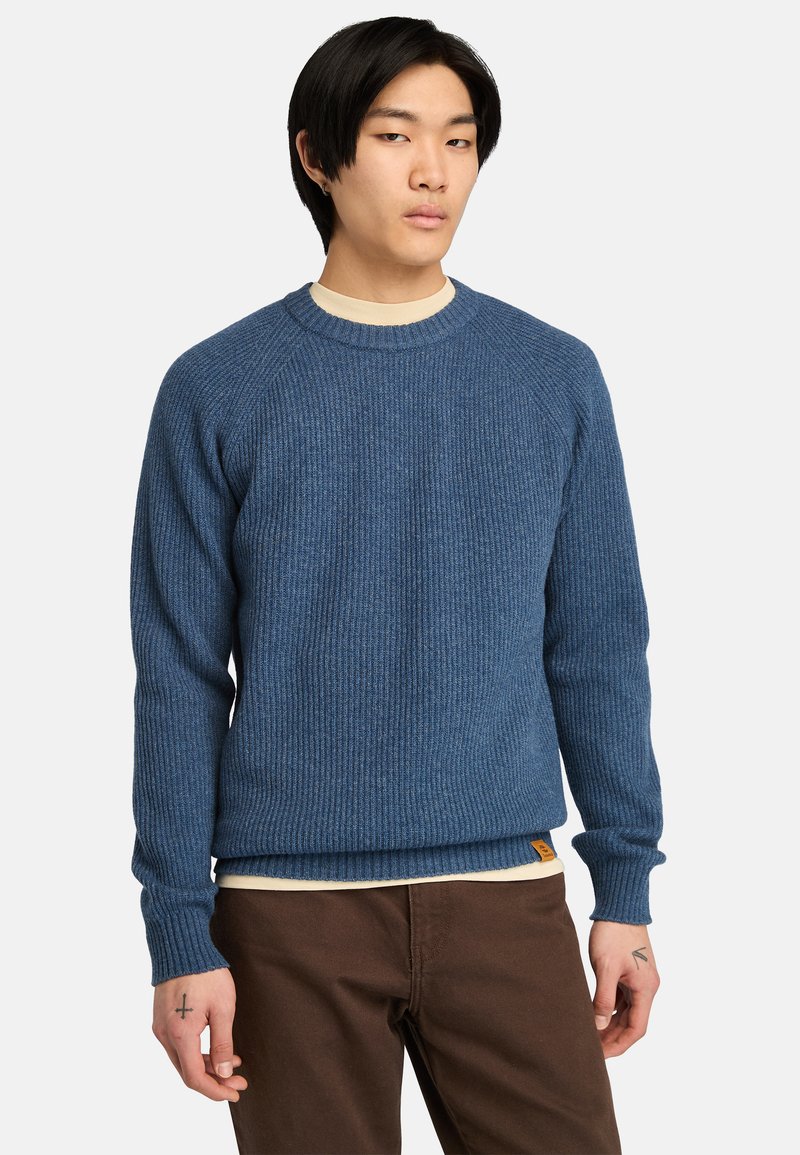 Blå ribbad tröja med rund halsringning och långa raglanärmar, tillsammans med en krämfärgad undertröja och bruna byxor.