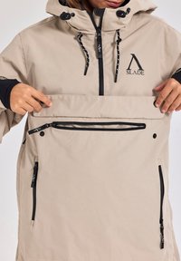 Beige waterdichte jas met een capuchon, zwarte rits en trekkoorden, voorzak en logo-detail. Heeft een gladde, textuurlijke afwerking.