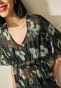 Femme portant un blouse noire transparente à motifs floraux avec des rayures verticales vertes et argentées, à manches courtes et ceinture à la taille, souriant avec du rouge à lèvres rouge.