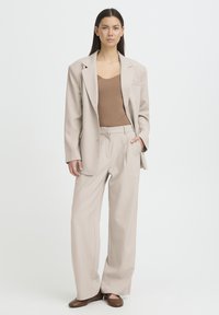 Veste beige sur mesure avec un col cranté, associée à un pantalon taille haute assorti et un haut marron ajusté. Ballerines marron texturées.