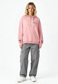 Felpa con cappuccio rosa e un tascone frontale, decorata con una grafica nera a croce; abbinata a pantaloni cargo grigi con tasche a pattina e polsini elasticizzati.