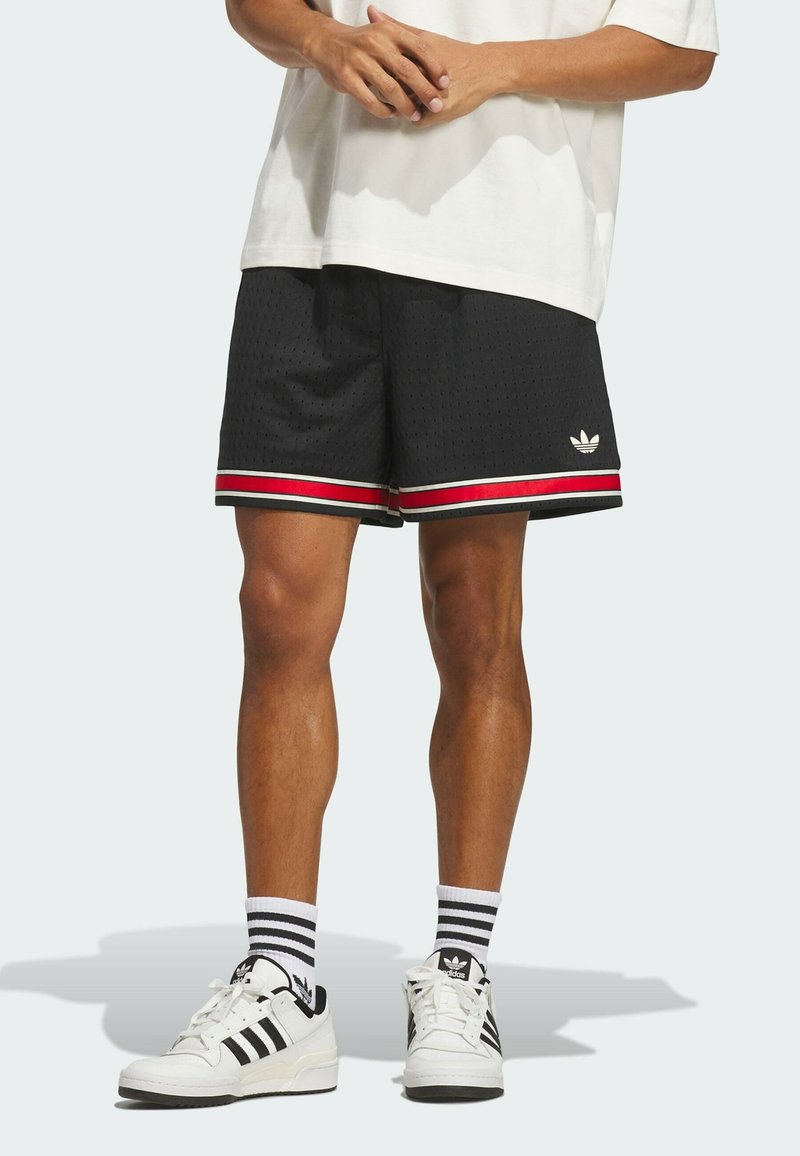 Person trägt schwarze Mesh-Shorts mit roten und weißen Streifen, weiße Adidas-Sneaker und weiße Socken mit schwarzen Streifen, steht vor neutralem Hintergrund.