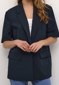 Blazer oscuro en color marino con mangas cortas, solapas muestradas y dos bolsillos frontales. Lleva encima una camisa blanca y está combinado con jeans azules.