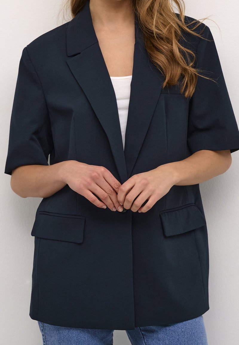 Blazer oscuro en color marino con mangas cortas, solapas muestradas y dos bolsillos frontales. Lleva encima una camisa blanca y está combinado con jeans azules.