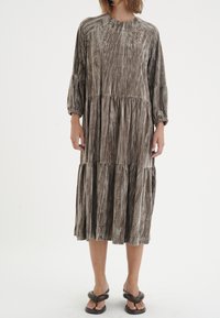 Kvinna klädd i en långärmad, tierad midiklänning i en strukturerad gråbrun tyg och svarta vadderade flip-flop-sandaler, stående mot en vit bakgrund.