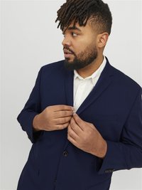 Navy blue blazer with a textured fabric; single-breasted design, button closure, and a chest pocket. Worn over a white shirt.