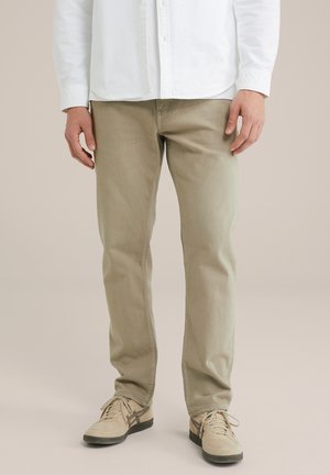 Man draagt beige broek, beige sneakers met zwarte zolen en een wit overhemd en staat voor een effen achtergrond.