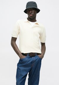 Κρεμ πλεκτό πόλο πουκάμισο με γιακά και τσέπη, συνδυασμένο με μπλε τζιν. Μαύρο καπέλο τύπου bucket και καφέ ζώνη ολοκληρώνουν την εμφάνιση.