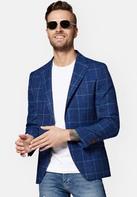 Niebieska marynarka w kratę o dopasowanym kroju, z delikatną teksturą, klapami w stylu notched oraz brązowymi guzikami. Noszona na białym t-shircie.