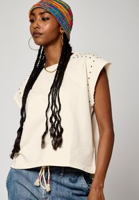 Crop top en crème à manches courtes avec des accents d'épaules cloutés et un ourlet à cordon. Associé à un denim bleu clair. Bonnet en tricot coloré sur la tête.