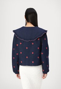 Ναυτικό τζάκετ από τζιν με μεγάλο ριχτό γιακά, κεντημένα με μικρές κόκκινες καρδιές και μακριά μανίκια. Διαθέτει ελαστικές μανσέτες.