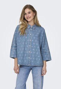 Svijetloplava traper jakna s ovratnikom, prevelikim rukavima i uzorkom srca. Zakopčava se s dugmadima sprijeda.