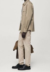 Chemise ample beige clair avec manches retroussées, associée à un pantalon crème et des chaussures en cuir noir. Veste en daim marron accrochée à proximité.