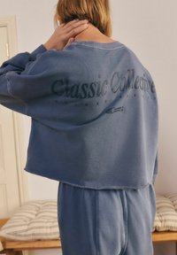 Przycięta, niebieska bluza z teksturowanej tkaniny. Na plecach ma napis "Classic Collective" w ciemnym kolorze.