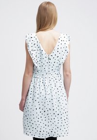 Femme portant une robe blanche à mi-longueur ornée de pois noirs irréguliers, avec des épaules volantées et une fermeture éclair visible dans le dos, faisant face à l'opposé de la caméra.