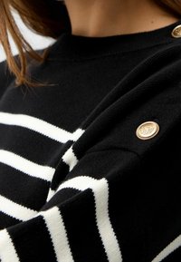 Pull noir avec des rayures blanches, col côtelé et détail de bouton doré sur l'épaule. Texture douce, vue en gros plan du tissu.