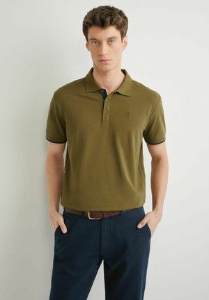 Mężczyzna w oliwkowej koszulce polo wpuszczonej w granatowe spodnie z brązowym plecionym paskiem, stojący na tle jasnej, jednolitej ściany.