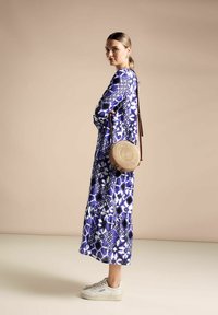 Lång blå klänning med geometriska mönster, graderad fåll, i kombination med en rund vävd väska och vita sneakers. Slät textur.