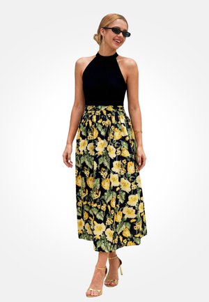 Kobieta w czarnym topie na ramiączkach, żółtej kwiecistej spódnicy midi, czarnych okularach przeciwsłonecznych i złotych sandałach na wysokim obcasie, uśmiechająca się i stojąca na tle białego tła.