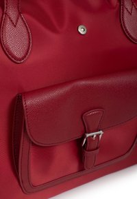 Rode handtas gemaakt van gestructureerde stof en leer. Beschikt over een klepzak met een zilveren gesp en een knoopsluiting. Over het algemeen een gladde afwerking.