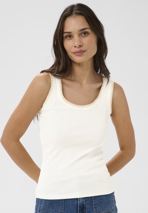 Ung kvinde med langt mørkt hår iført en hvid ærmeløs ribstrikket top med hæklet kant, stående foran en ensfarvet lys grå baggrund.