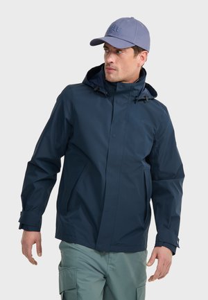 Navyblauer wasserdichter Mantel mit Zugkordelkapuze, Druckknöpfen und Seitentaschen. Getragen mit hellgrünen Cargo-Hosen und einer hellblauen Mütze.