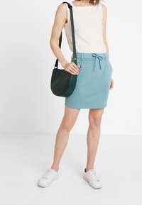 Niebieska rozciągliwa spódnica z wiązanym pasem, zestawiona z białym bezrękawnikiem i ciemnozieloną torbą na ramię. Całość dopełniają białe trampki.