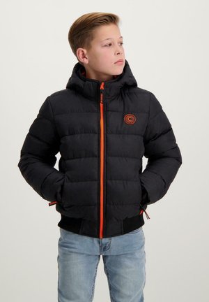 Boy wearing a black padded jacket with an orange zipper and logo, standing confidently with his hands in his pockets against a white background.
