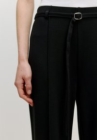 Schwarze maßgeschneiderte Hosen aus strukturiertem Material, mit einem Gürtelschlaufen mit einer silbernen Schnalle und einem hängenden Bindegürtel-Detail an der Taille.