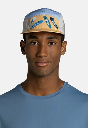 Junger Mann, der eine hellblaue und beige Kappe mit blauen Kajakmotiven und ein blaues Rundhals-T-Shirt trägt, schaut geradeaus mit neutralem Gesichtsausdruck.