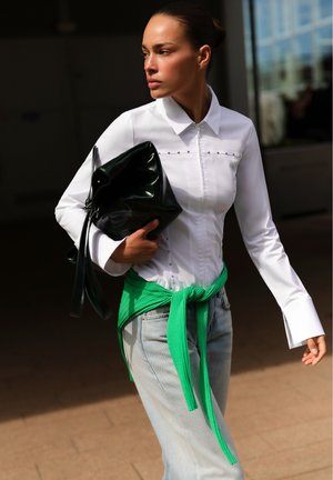 Femme portant une chemise blanche zippée et un jean clair avec un pull vert noué autour de la taille, tenant un grand sac noir.