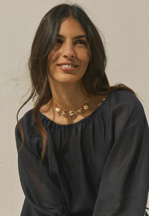 Frau mit langen dunklen Haaren, die ein schwarzes schulterfreies Oberteil und eine Halskette mit goldenen Muschel- und Seesternanhängern trägt, lächelt sanft.