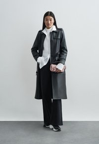 Svart lær trenchcoat med knappelukking, kombinert med en hvit skjorte og brede svarte bukser. Tilbehøret inkluderer en liten rosa håndveske.