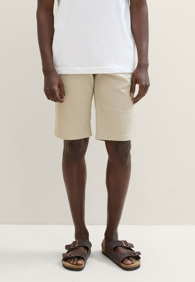 Shorts beiges en coton, à mi-genou avec une coupe droite. Associés à des sandales marron foncé avec des boucles réglables.